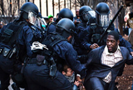five heavily armored and armed police officers pressing a black man in a business suit to his knees