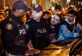 two police officers talking at the back of a car, surrounded by youths